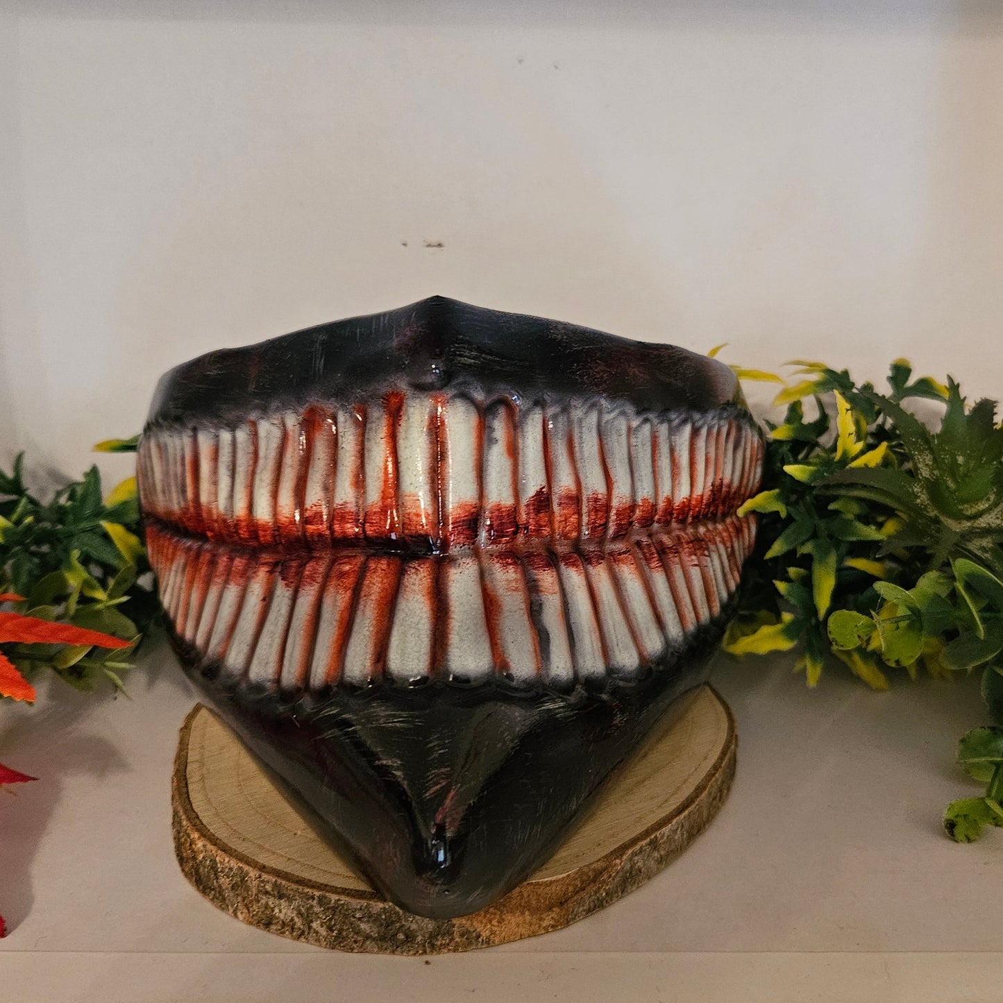 Ein auf einem Holzsockel platziertes Cosplay-Requisit in Form eines großen, gruseligen Gebisses mit zahlreichen Zähnen, die mit roter Farbe blutähnlich bemalt sind. Umgeben von künstlichen Pflanzen, darunter grüne Blätter und rote Ahornblätter. Ideal für ein Horror-Cosplay oder eine Halloween-Dekoration.