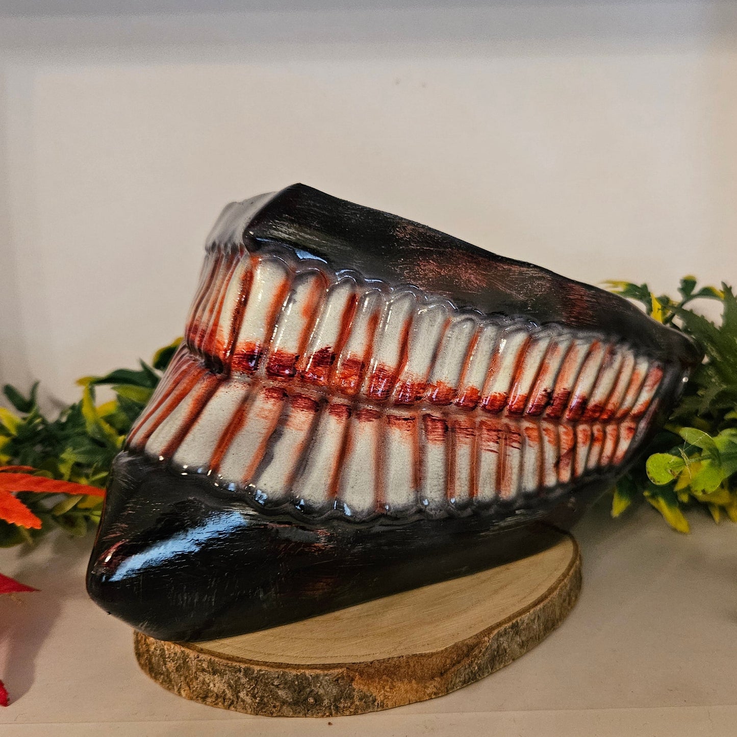 Ein auf einem Holzsockel platziertes Cosplay-Requisit in Form eines großen, gruseligen Gebisses mit zahlreichen Zähnen, die mit roter Farbe blutähnlich bemalt sind. Umgeben von künstlichen Pflanzen, darunter grüne Blätter und rote Ahornblätter. Ideal für ein Horror-Cosplay oder eine Halloween-Dekoration.