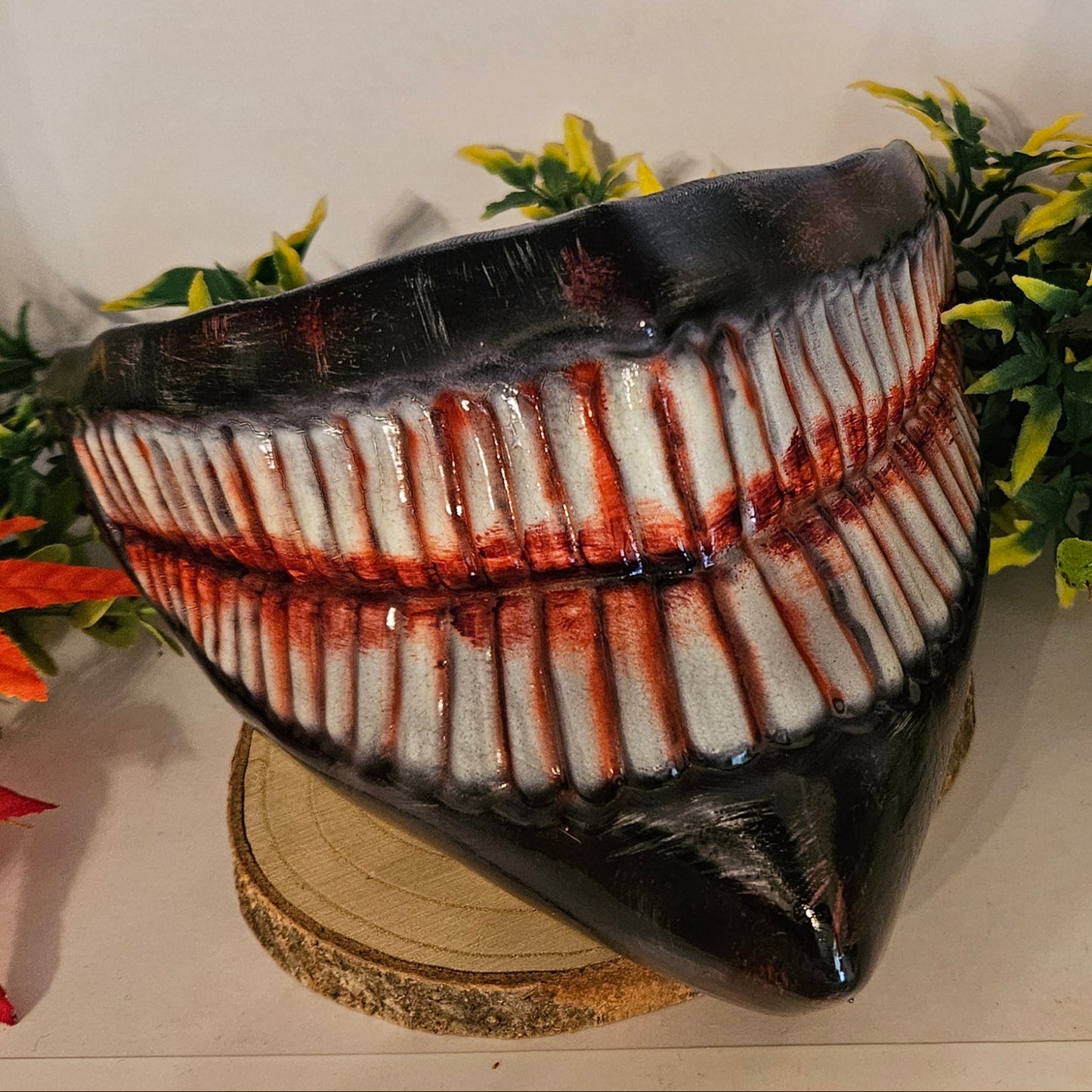 Ein auf einem Holzsockel platziertes Cosplay-Requisit in Form eines großen, gruseligen Gebisses mit zahlreichen Zähnen, die mit roter Farbe blutähnlich bemalt sind. Umgeben von künstlichen Pflanzen, darunter grüne Blätter und rote Ahornblätter. Ideal für ein Horror-Cosplay oder eine Halloween-Dekoration.