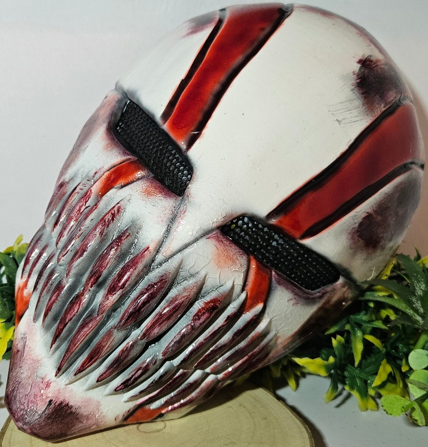 Eine detaillierte und kunstvoll gestaltete Maske mit einem Horror- oder Fantasy-Thema. Sie ist überwiegend weiß mit roten und schwarzen Akzenten, besitzt scharfe, bedrohliche Zähne und dunkle Augenschlitze. Die Maske steht auf einem Holzständer und ist umgeben von künstlichem grünen und gelben Laub, mit einem einzelnen roten und orangefarbenen Blatt im Vordergrund. Das Design ist eindrucksvoll und furchteinflößend, passend für Fans von Horror- oder Fantasy-Ästhetik.