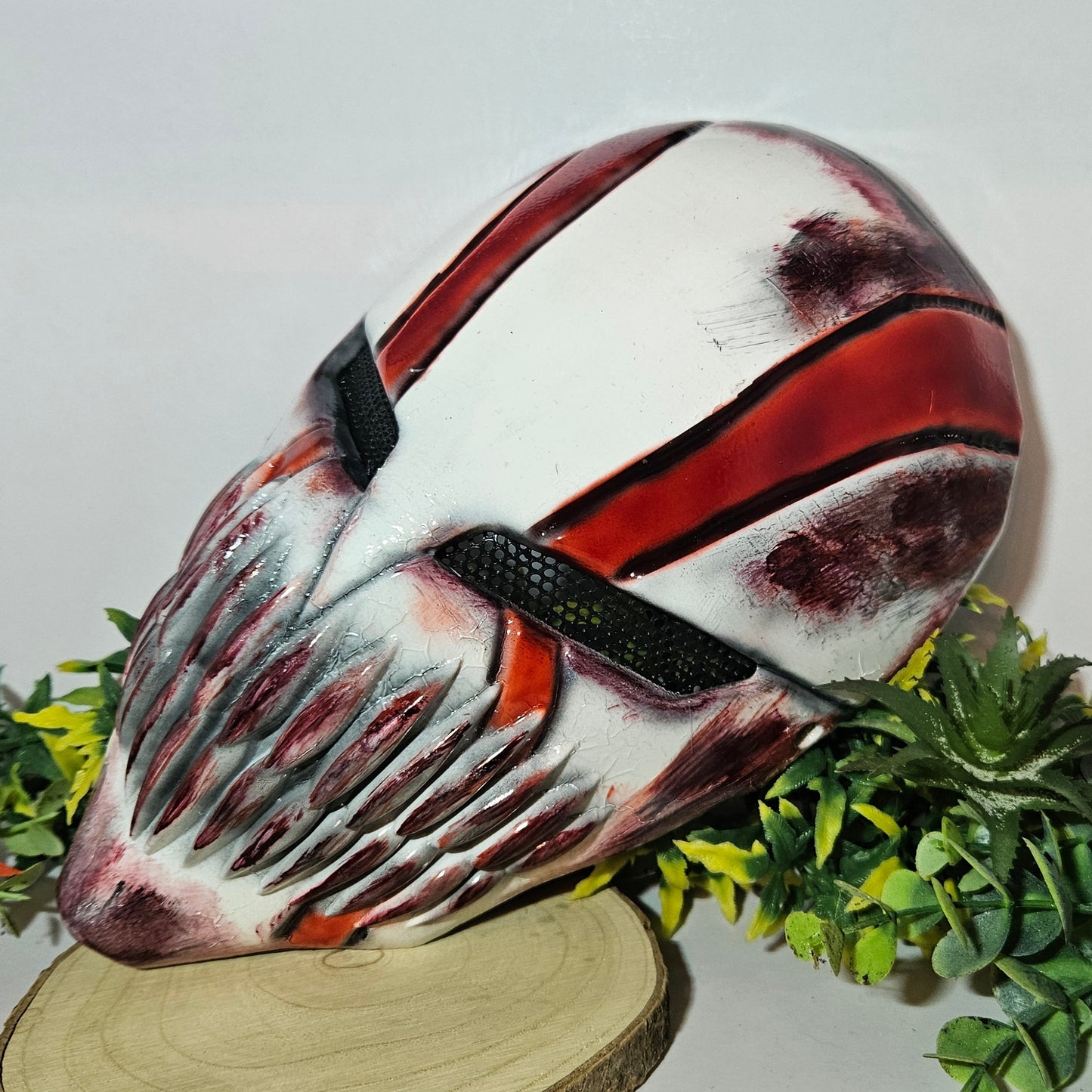 Eine detaillierte und kunstvoll gestaltete Maske mit einem Horror- oder Fantasy-Thema. Sie ist überwiegend weiß mit roten und schwarzen Akzenten, besitzt scharfe, bedrohliche Zähne und dunkle Augenschlitze. Die Maske steht auf einem Holzständer und ist umgeben von künstlichem grünen und gelben Laub, mit einem einzelnen roten und orangefarbenen Blatt im Vordergrund. Das Design ist eindrucksvoll und furchteinflößend, passend für Fans von Horror- oder Fantasy-Ästhetik.