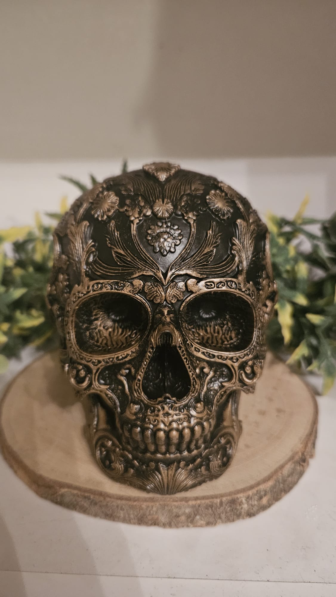 Handbemalte mystische Gothic Deko Figur in Form eines ornamentalen Skulls aus Kunstharz, ca. 10 cm groß, mit goldenen Verzierungen auf dunklem Grund, präsentiert auf einer Holzplatte mit dekorativen Pflanzen im Hintergrund