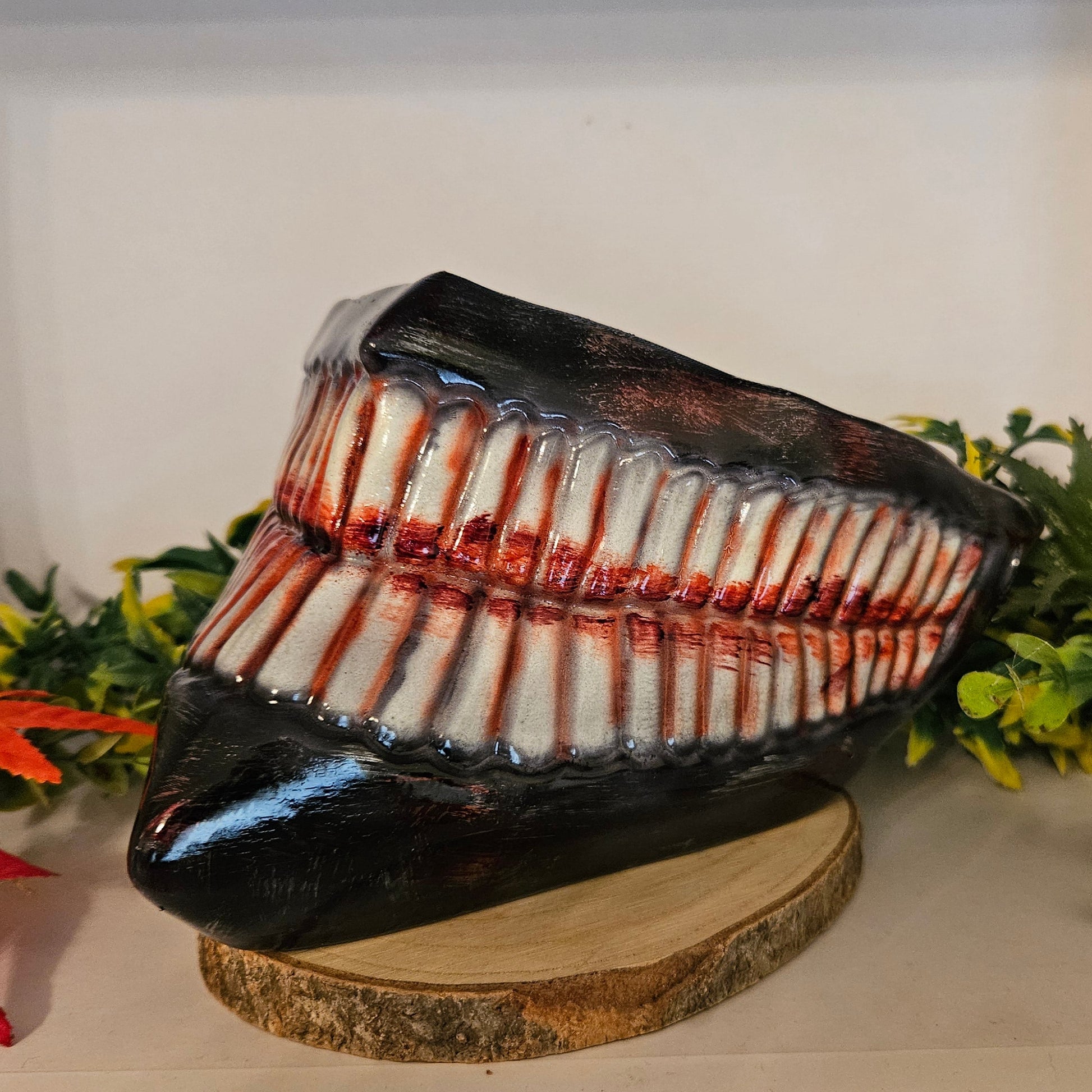  Ein auf einem Holzsockel platziertes Cosplay-Requisit in Form eines großen, gruseligen Gebisses mit zahlreichen Zähnen, die mit roter Farbe blutähnlich bemalt sind. Umgeben von künstlichen Pflanzen, darunter grüne Blätter und rote Ahornblätter. Ideal für ein Horror-Cosplay oder eine Halloween-Dekoration.