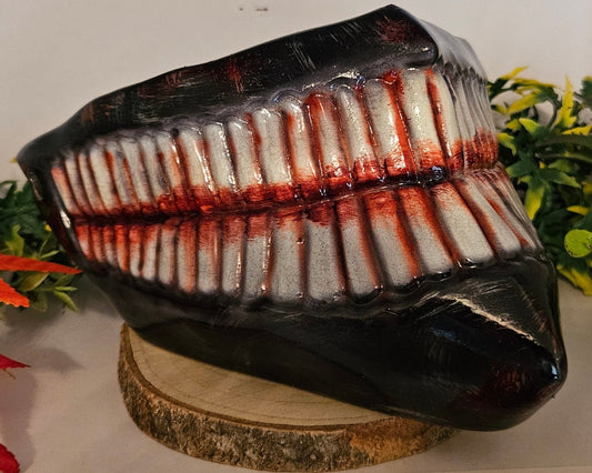  Ein auf einem Holzsockel platziertes Cosplay-Requisit in Form eines großen, gruseligen Gebisses mit zahlreichen Zähnen, die mit roter Farbe blutähnlich bemalt sind. Umgeben von künstlichen Pflanzen, darunter grüne Blätter und rote Ahornblätter. Ideal für ein Horror-Cosplay oder eine Halloween-Dekoration.