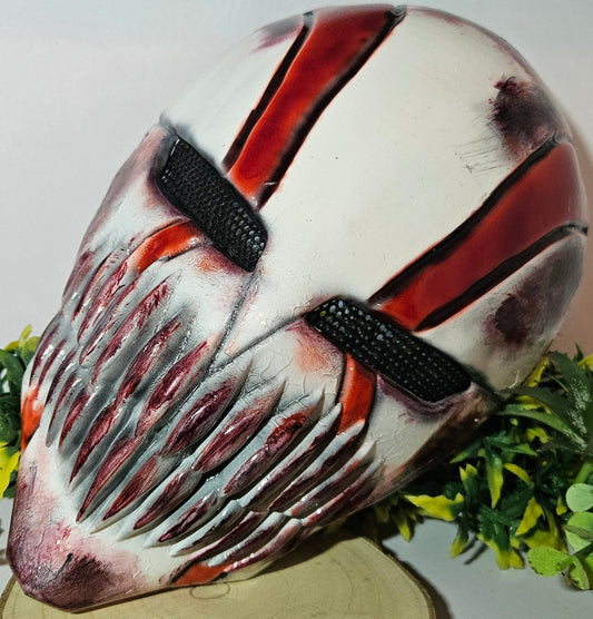Eine detaillierte und kunstvoll gestaltete Maske mit einem Horror- oder Fantasy-Thema. Sie ist überwiegend weiß mit roten und schwarzen Akzenten, besitzt scharfe, bedrohliche Zähne und dunkle Augenschlitze. Die Maske steht auf einem Holzständer und ist umgeben von künstlichem grünen und gelben Laub, mit einem einzelnen roten und orangefarbenen Blatt im Vordergrund. Das Design ist eindrucksvoll und furchteinflößend, passend für Fans von Horror- oder Fantasy-Ästhetik.